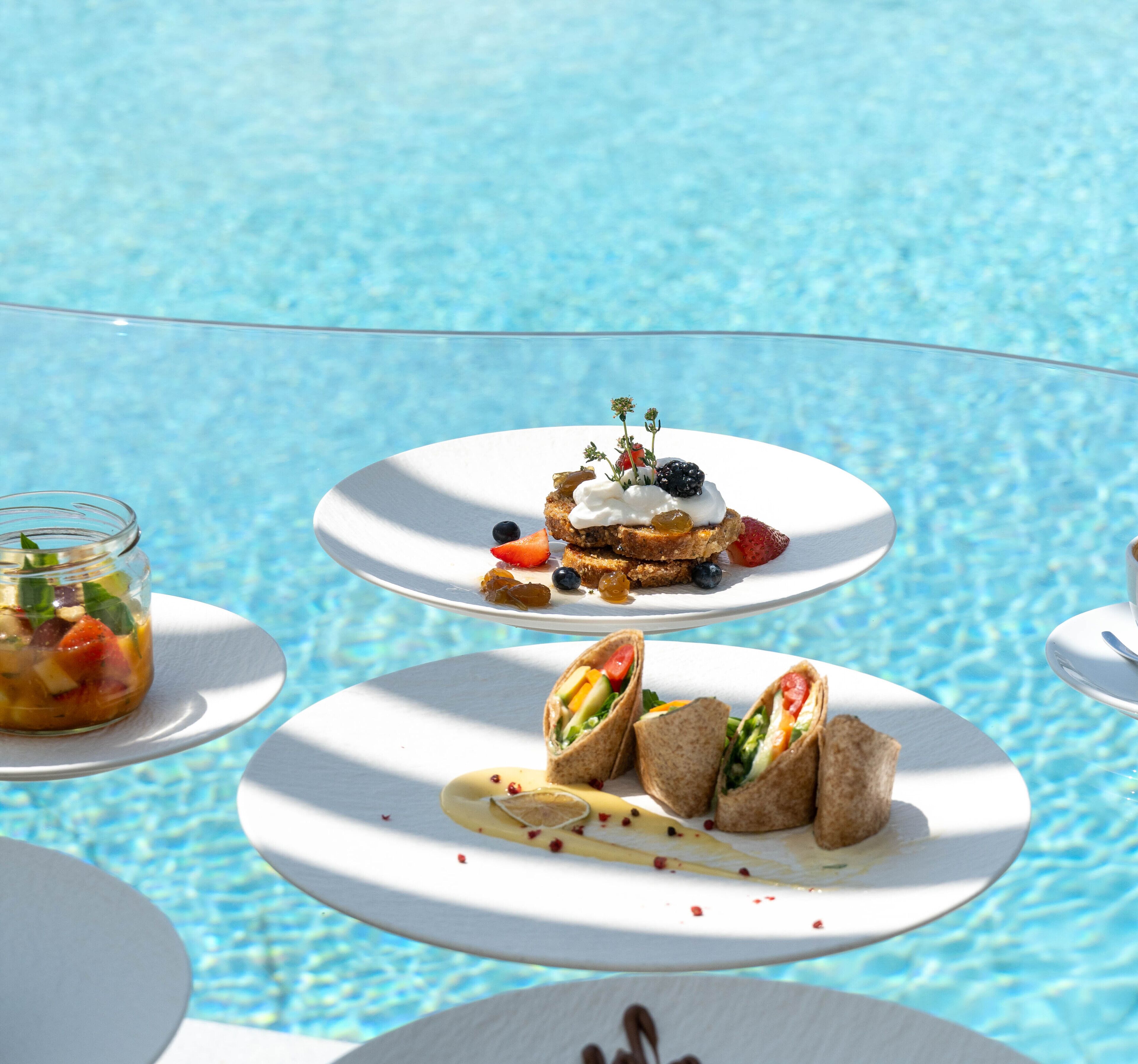 A poolside dining area with white plates of food, including sandwiches and a dessert with berries and cream.