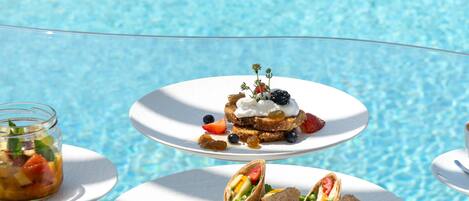 A poolside dining area with white plates of food, including sandwiches and a dessert with berries and cream.