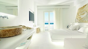 A modern hotel room with a large stone sink, a flat-screen TV, a bed, and a balcony with a view of the sea.