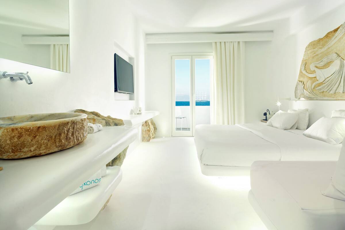 Une chambre d’hôtel moderne dotée d’un grand lavabo en pierre, d’un téléviseur à écran plat, d’un lit et d’un balcon offrant une vue sur la mer.