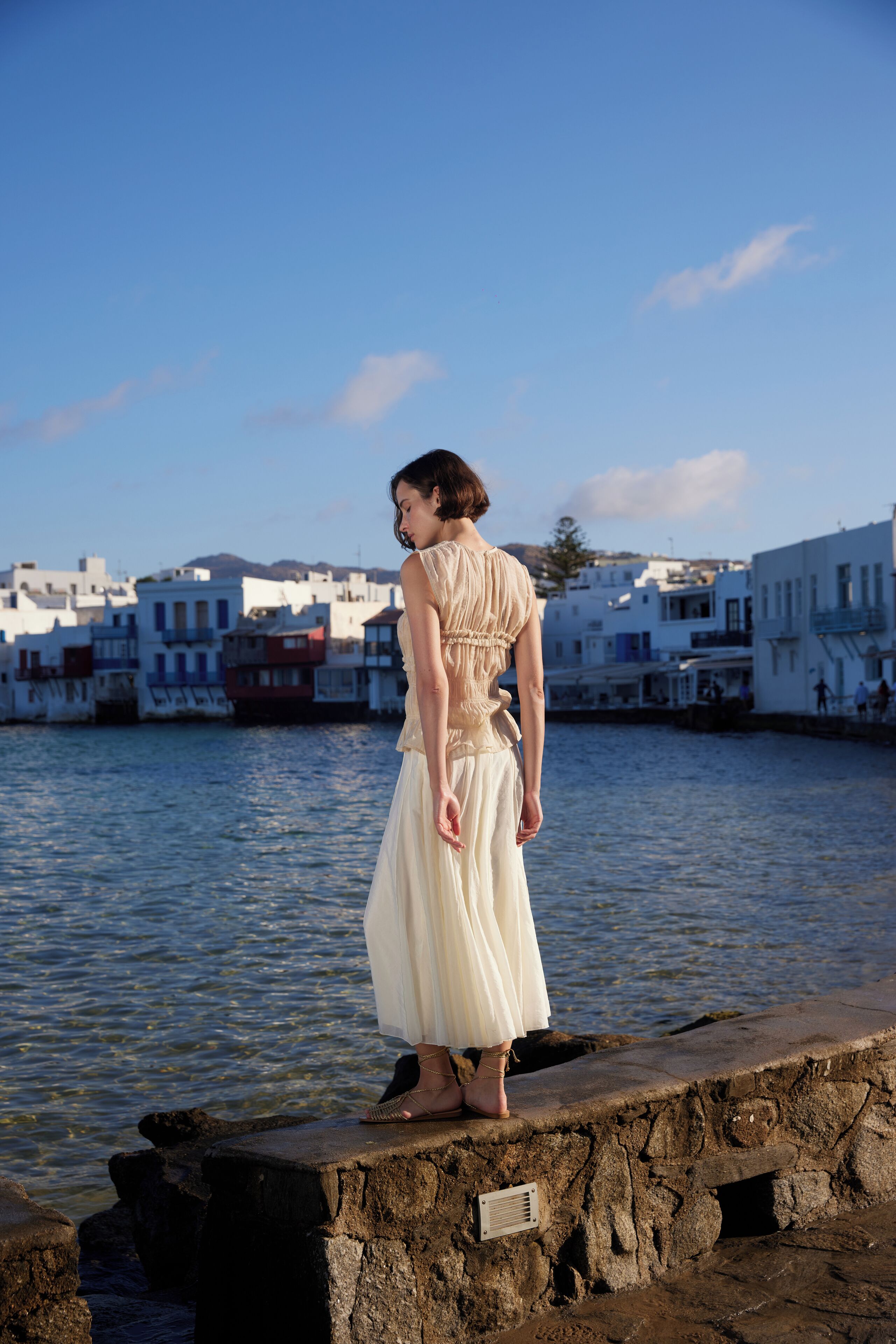 A woman standing on a stone platform by the water, overlooking a coastal town with white buildings.