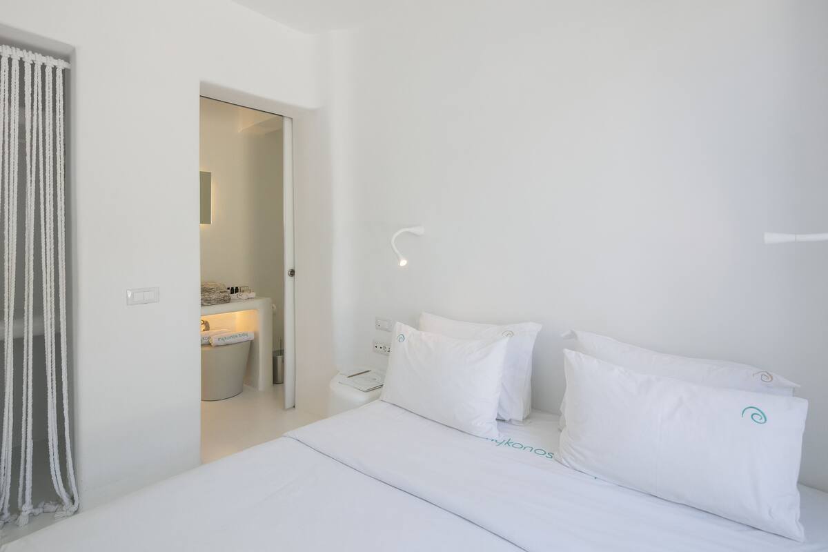 Une chambre d’hôtel au style minimaliste, avec un lit soigneusement fait, une table de chevet avec une lampe et une salle de bain visible à travers une porte ouverte.