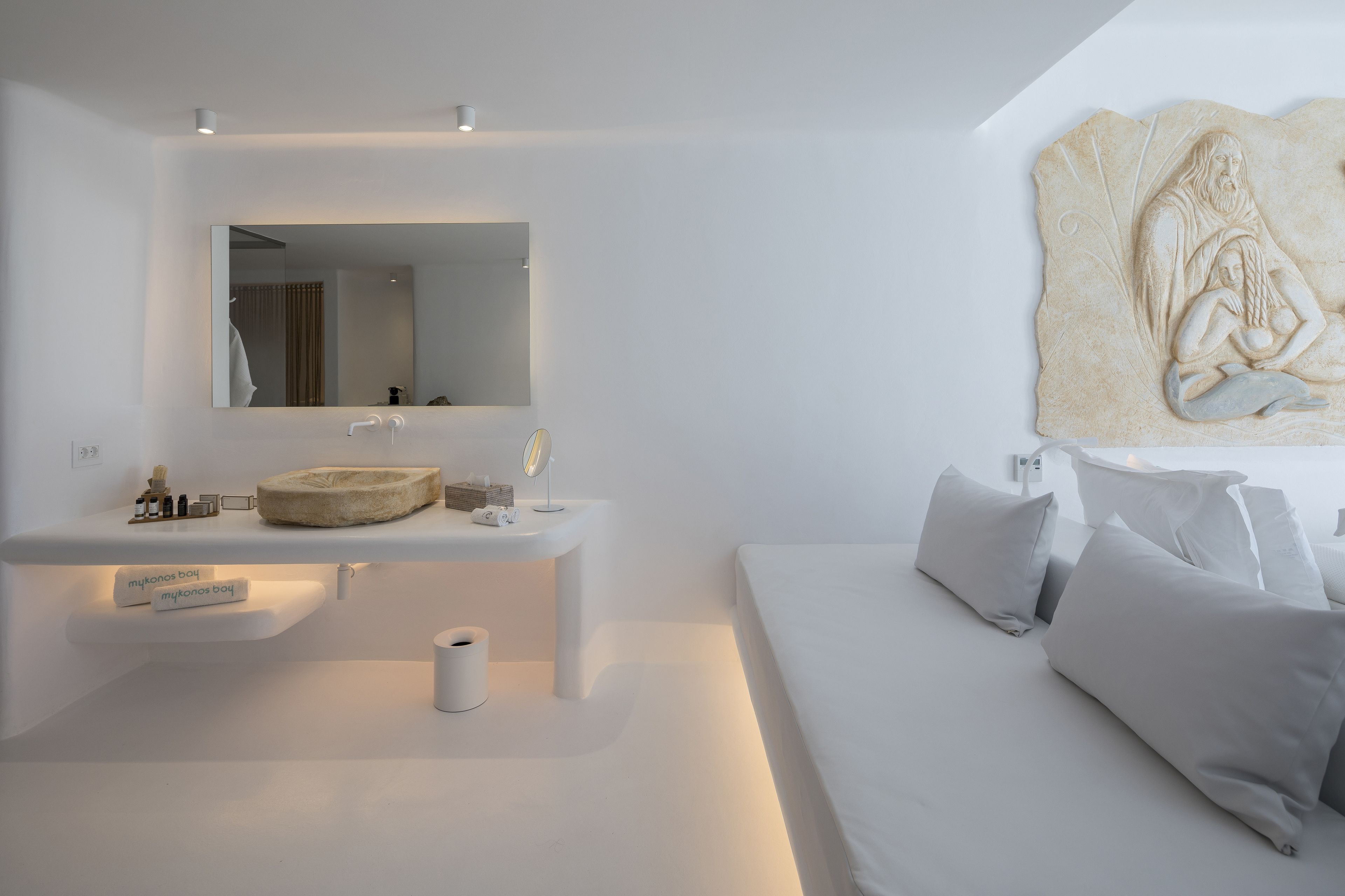 A modern, minimalist bathroom with a large mirror, a round sink, and a toilet. There is a bed with white bedding and pillows in the adjacent room.