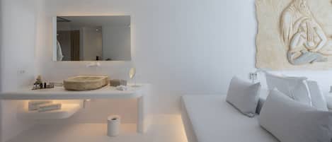 Ett modernt, minimalistiskt badrum med en stor spegel, ett runt handfat och en toalett. I det intilliggande rummet finns en säng med vita sängkläder och kuddar.