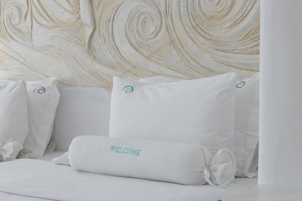 Un lit avec une literie et des oreillers blancs, un coussin orné de l’inscription « BIENVENUE », et un mur à texture en arrière-plan.