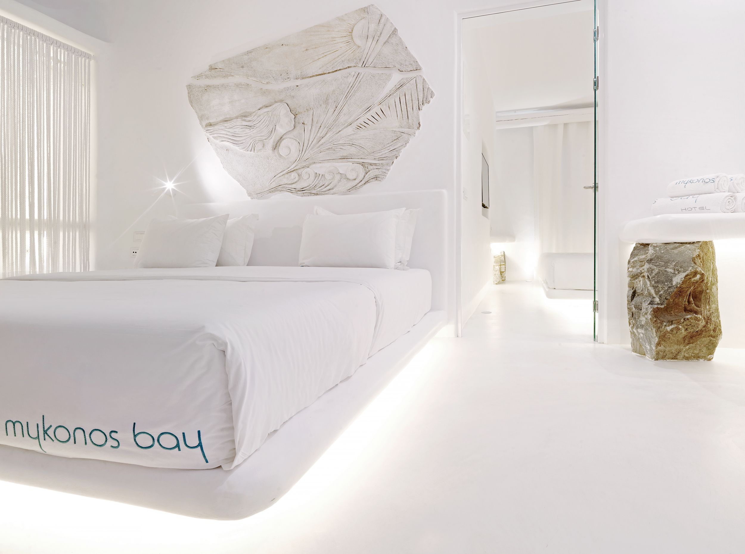 A modern hotel room with a large bed, a stone accent piece, and a view of the hallway.
