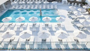 A pool area with multiple white lounge chairs and umbrellas, a clear blue pool, and a stone wall in the background.