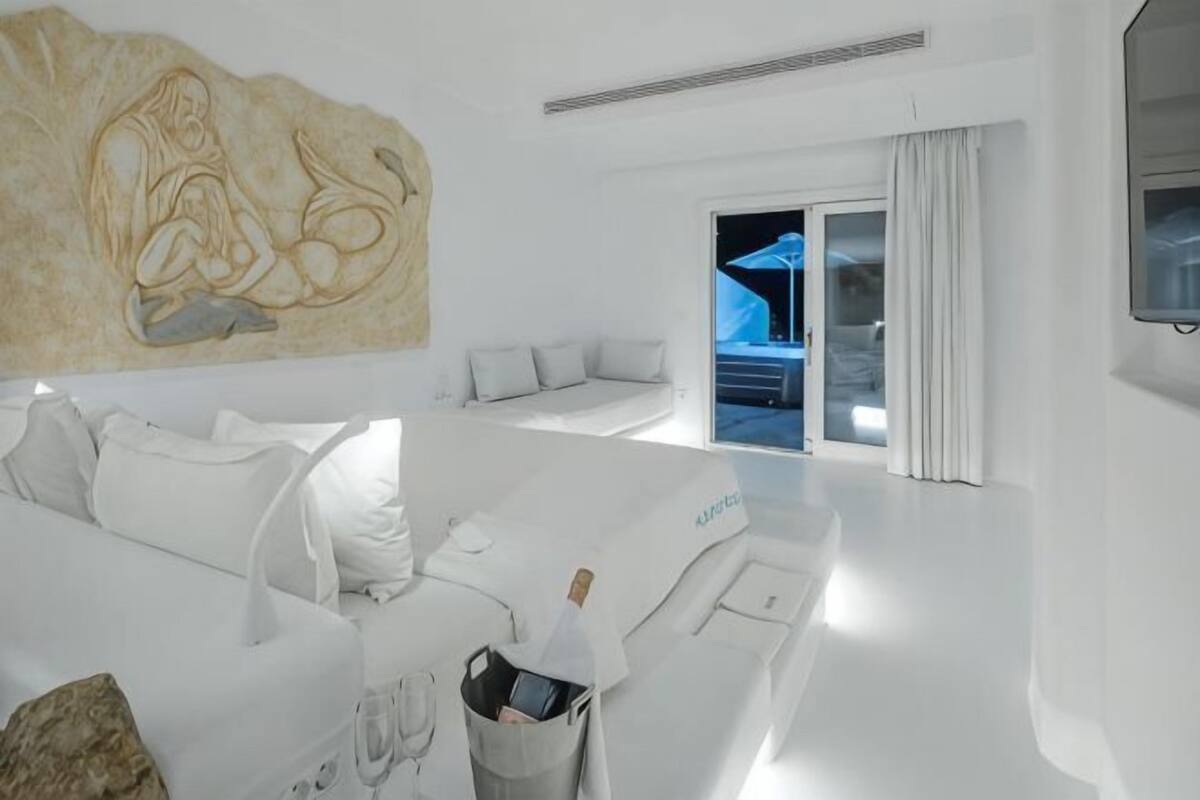 Une chambre d’hôtel moderne dotée d’un grand lit, d’un coin salon avec un canapé et des fauteuils, d’un téléviseur fixé au mur et d’une grande fenêtre avec des rideaux.