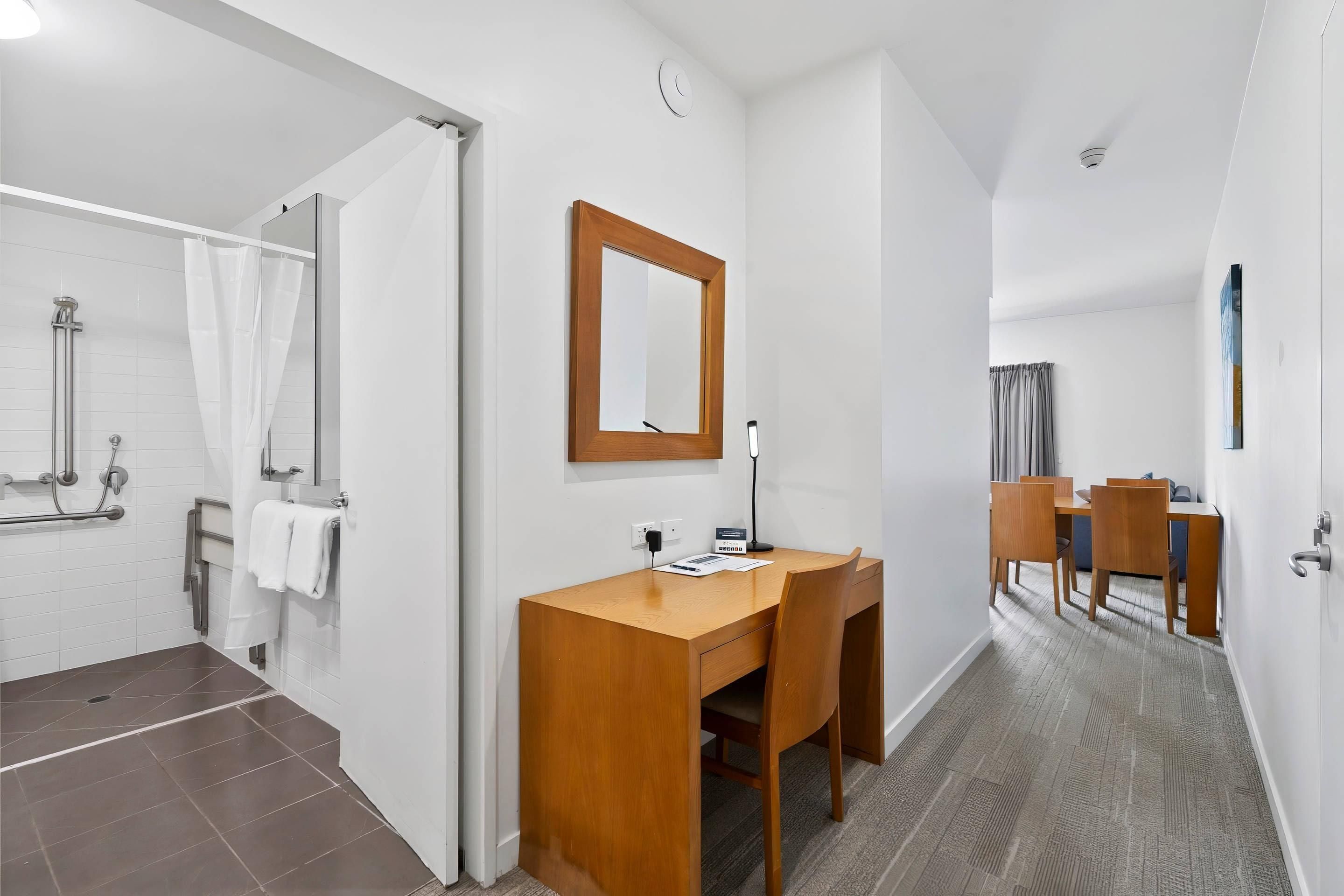 Habitación de hotel con ducha, un escritorio de madera, una silla, un espejo y un área de comedor con mesa y sillas.