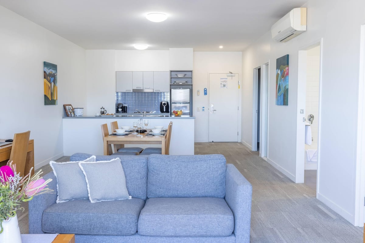 A modern living room with a grey sofa, a dining table with chairs, a kitchen area with white cabinets and appliances, and a hallway leading to a bathroom.