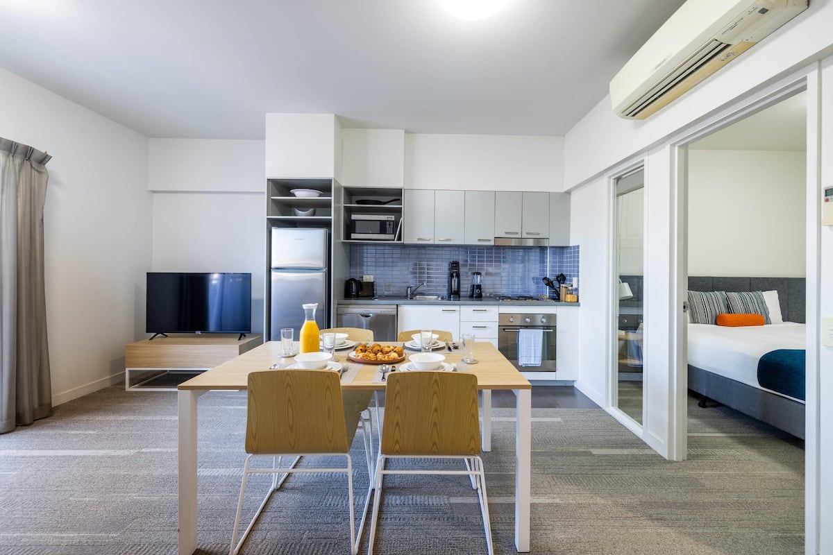 A modern hotel room with a dining area, kitchenette, and a bed.