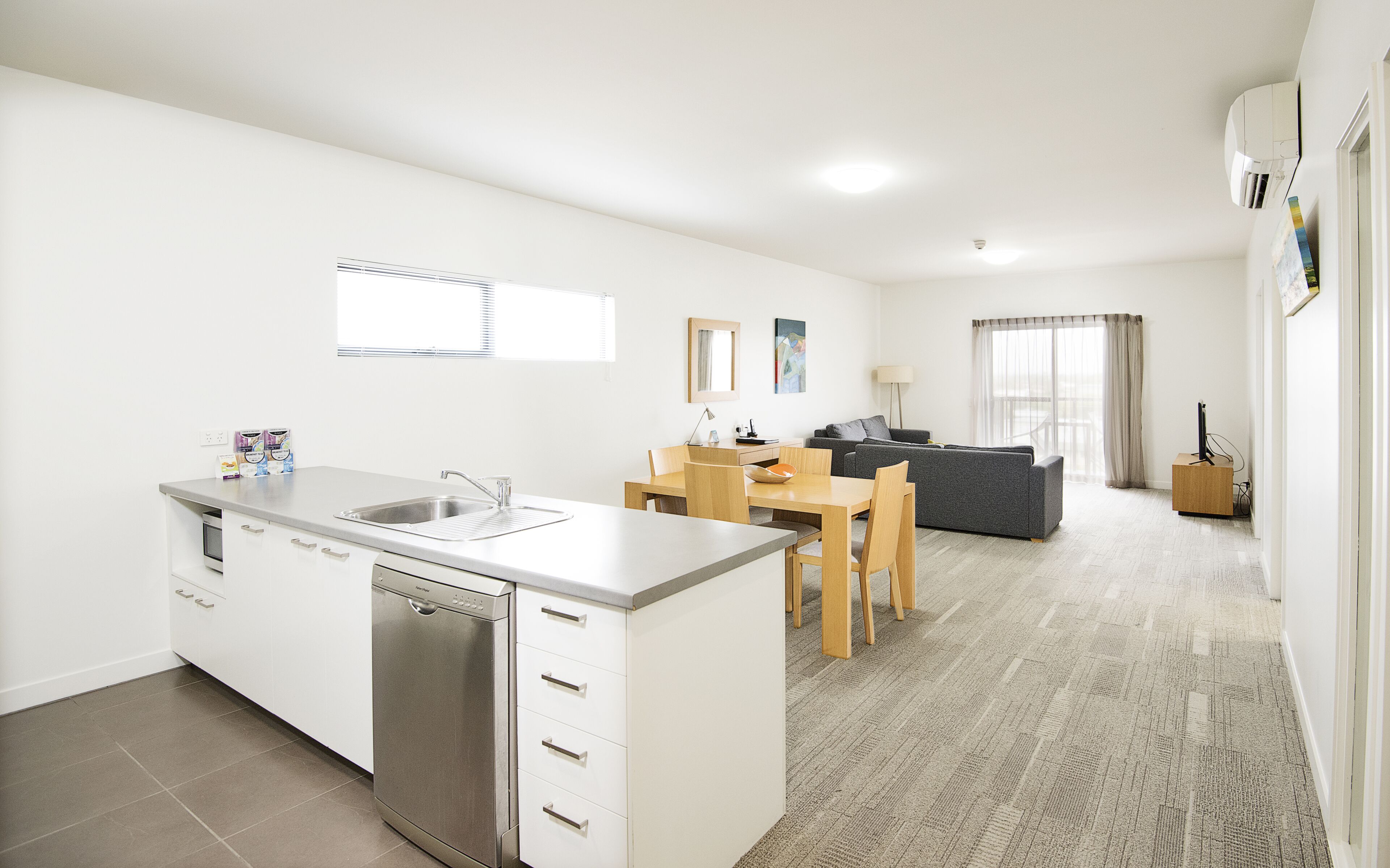 A modern kitchen with a dishwasher, a dining area with a table and chairs, a gray sofa, and a view of the outdoors through a window.