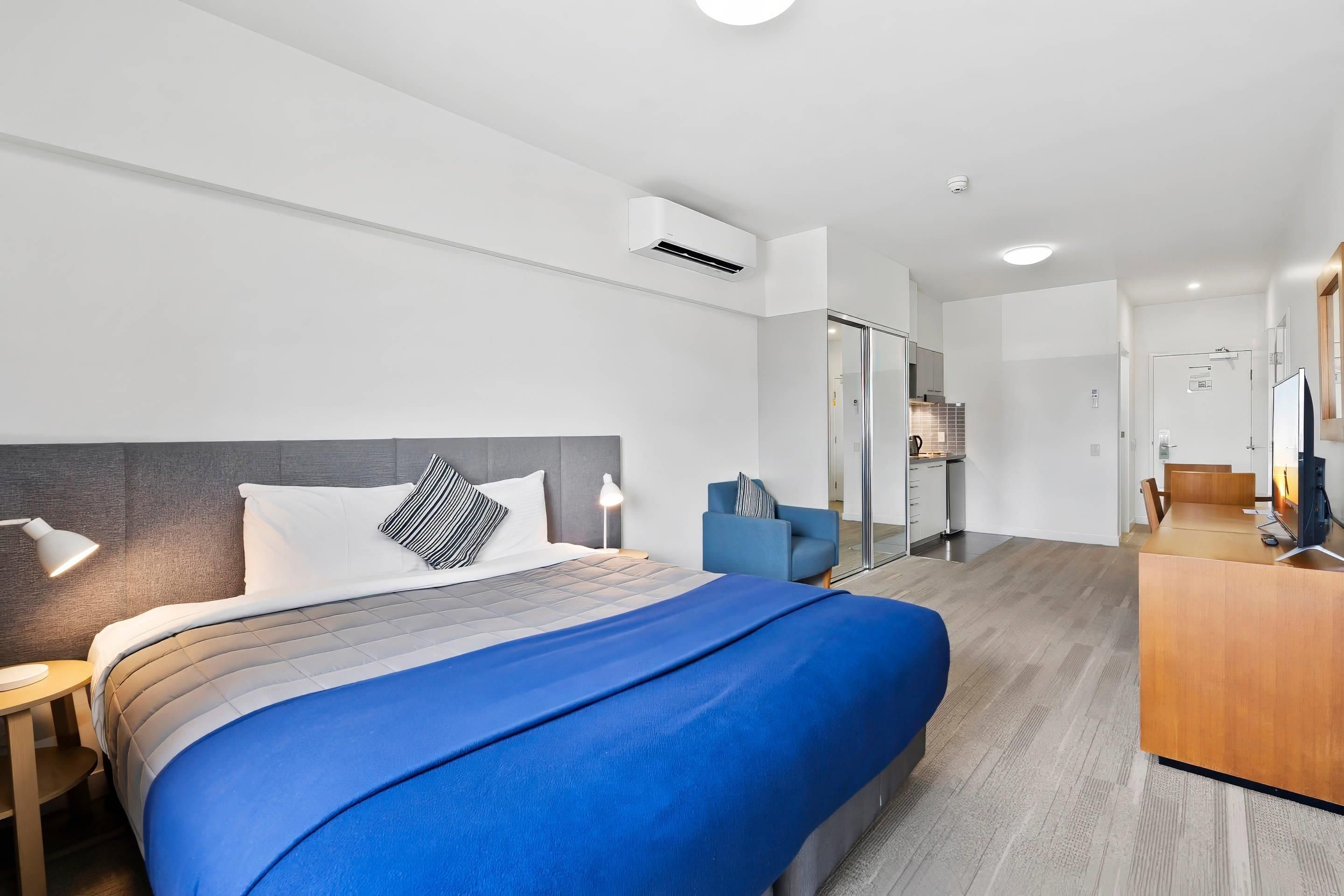 A modern hotel room with a bed, bedside table, lamp, and a view into the kitchen area.