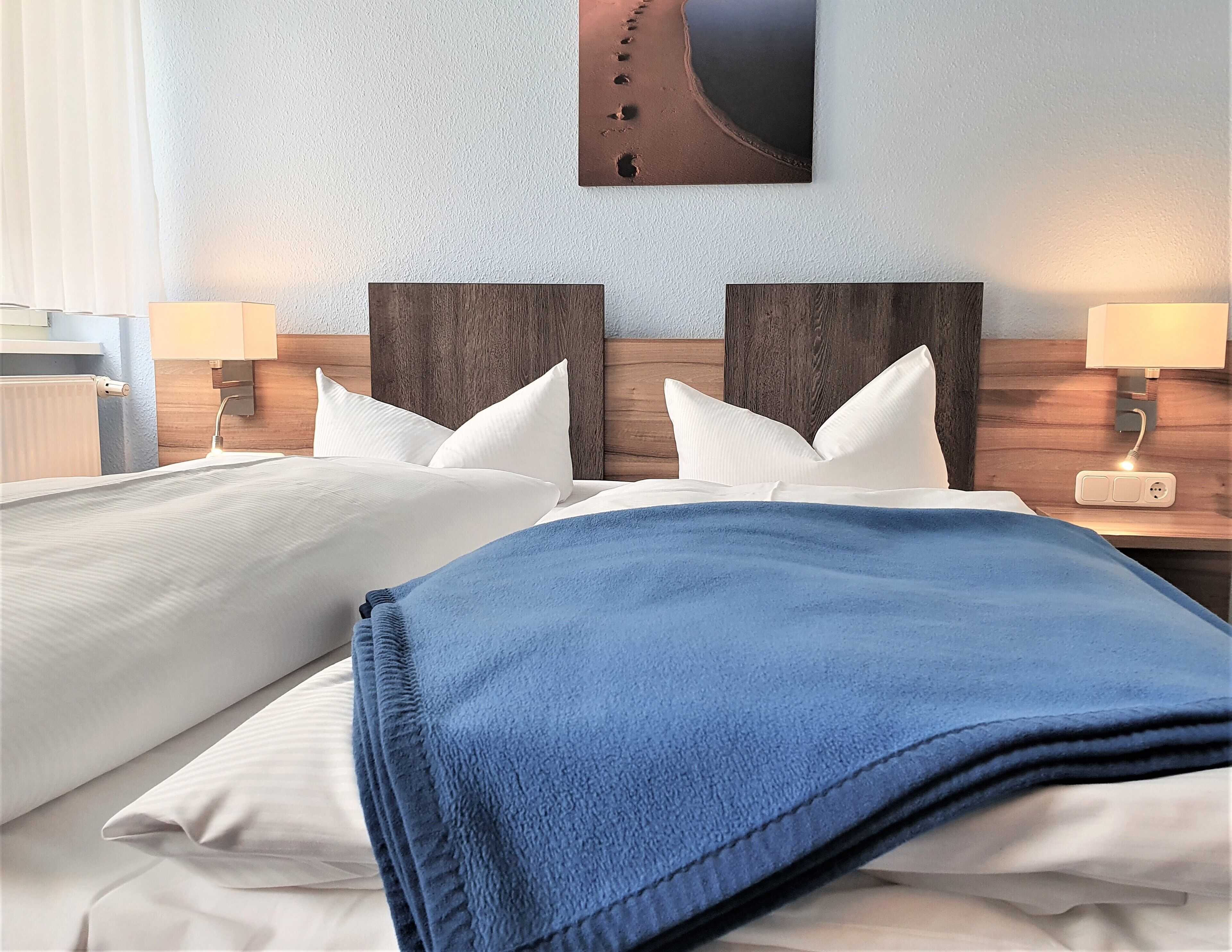 Ein Hotelzimmer mit zwei Betten, weißen Laken, einer blauen Decke, Holz-Kopfteilen und einem Bild mit einer Strandansicht.
