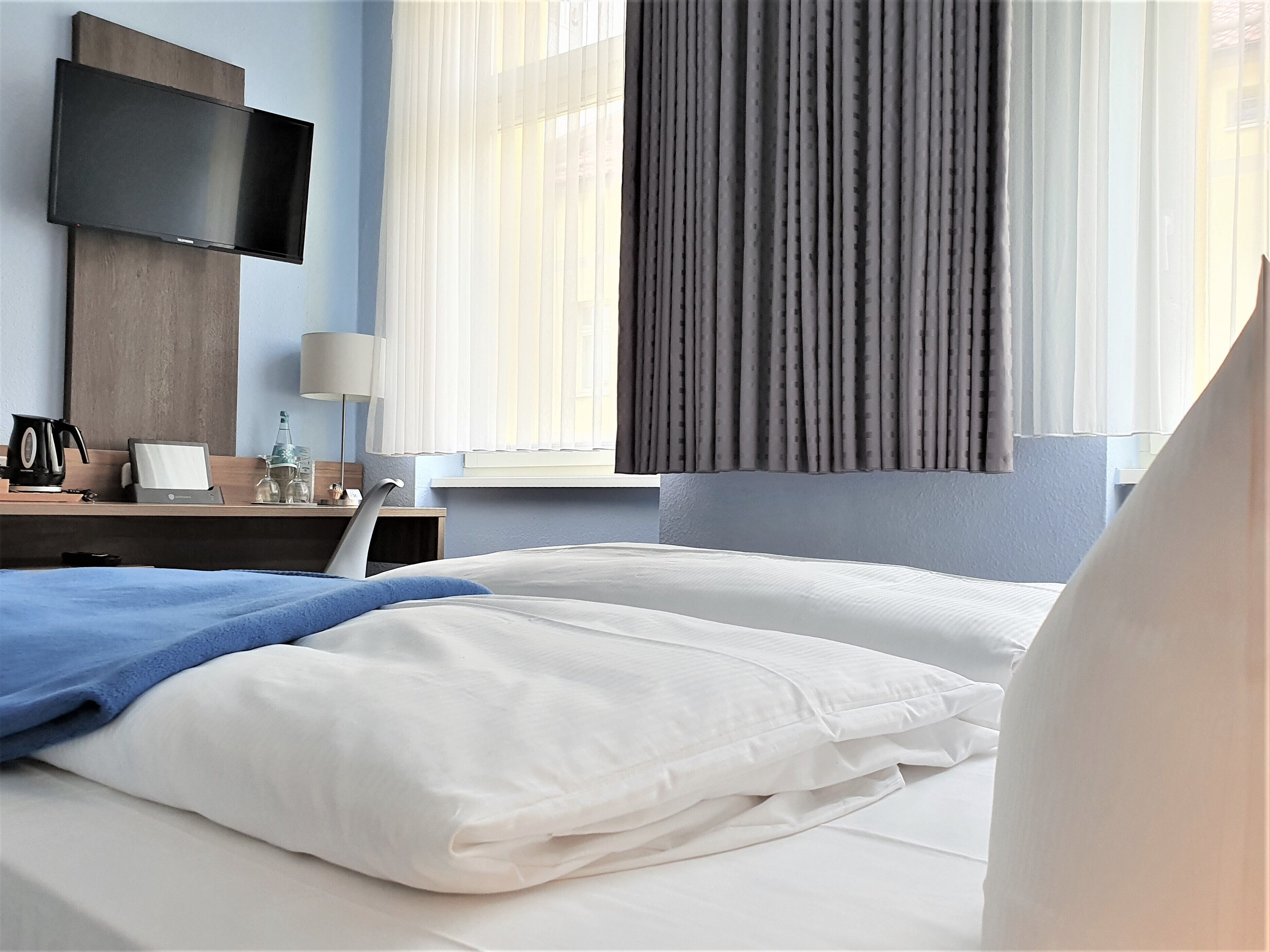 A hotel room with a bed, a desk with a lamp, a television, and a window with curtains.