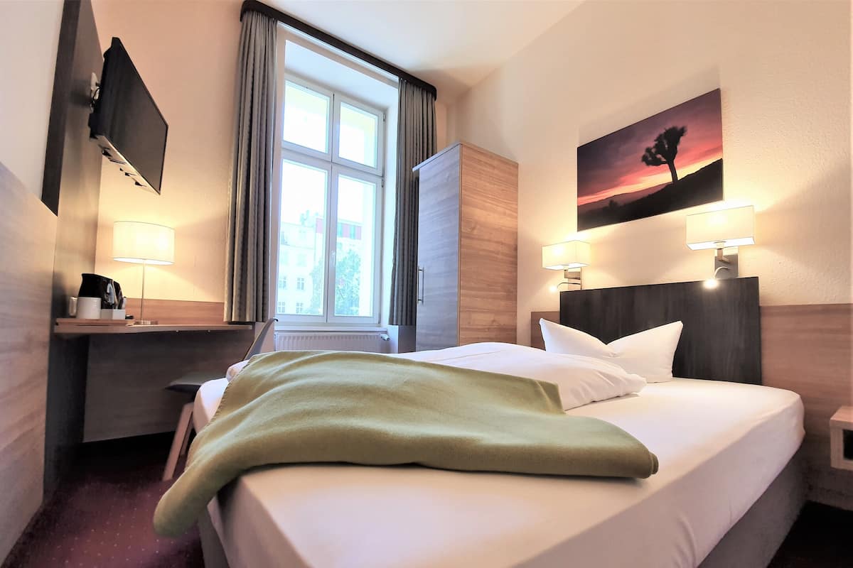 A hotel room with a bed, a desk, a lamp, a painting, and a window.