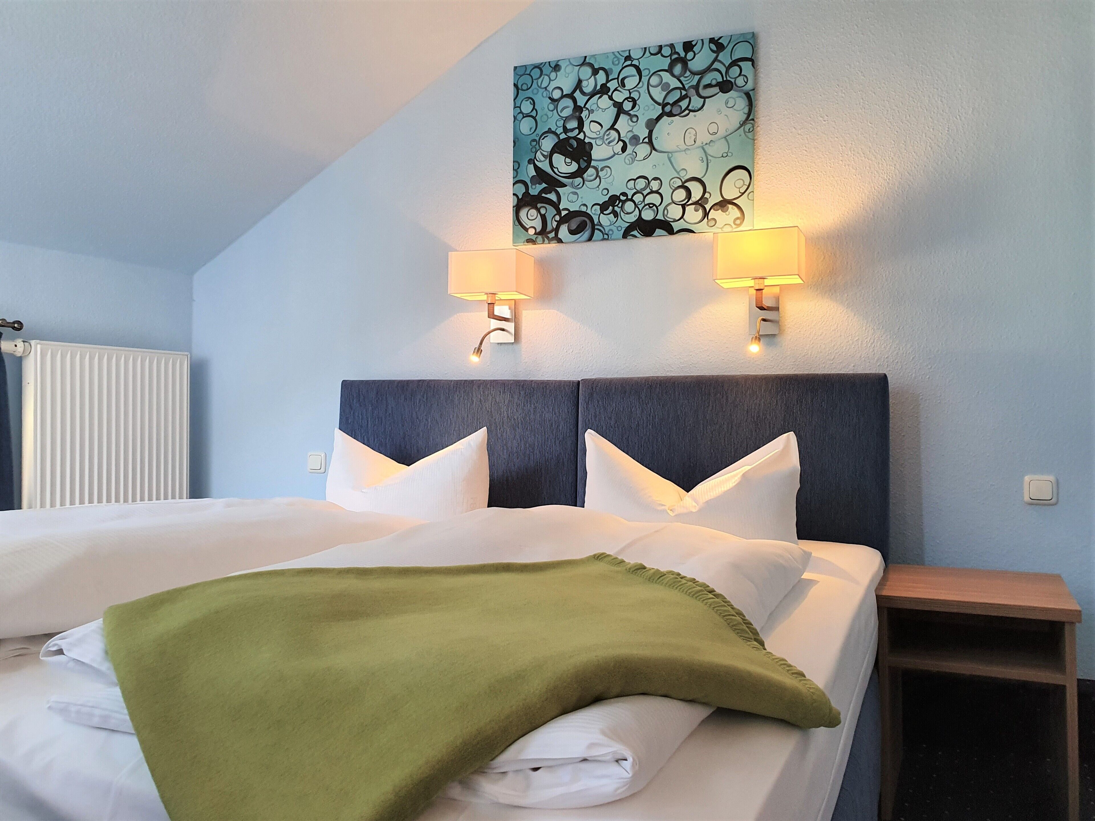 A neatly made bed with white linens and a green throw blanket, two wall-mounted lamps, a radiator, and a wall art piece above the bed.