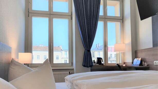 Habitación de hotel con dos camas, un escritorio con una computadora portátil, una cafetera y vistas a edificios a través de amplios ventanales.
