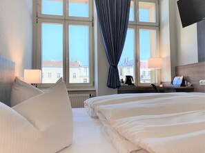 A hotel room with two beds, a desk with a laptop, a coffee maker, and a view of buildings through large windows. - Melarose Feng Shui Hotel (Berlin)