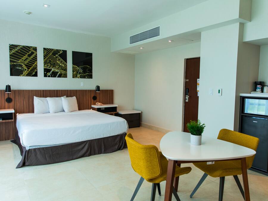 Habitación de hotel con cama, mesitas de noche, escritorio con silla, refrigerador pequeño y una obra de arte en la pared.