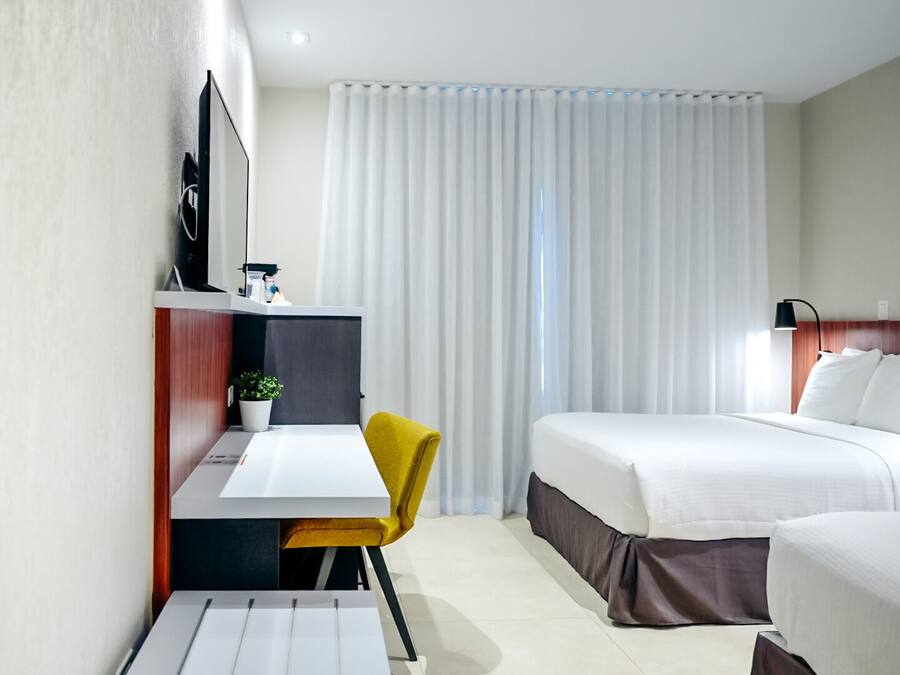 Habitación de hotel moderna con cama, escritorio, silla, televisor y ventana con cortinas.