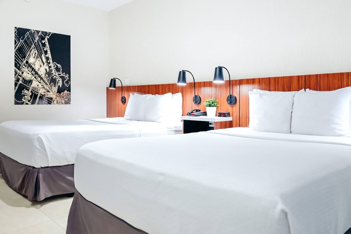 Ein Hotelzimmer mit zwei Betten, einem Holzkopfende, einem Nachttisch, einer Uhr und einer Wandleuchte.