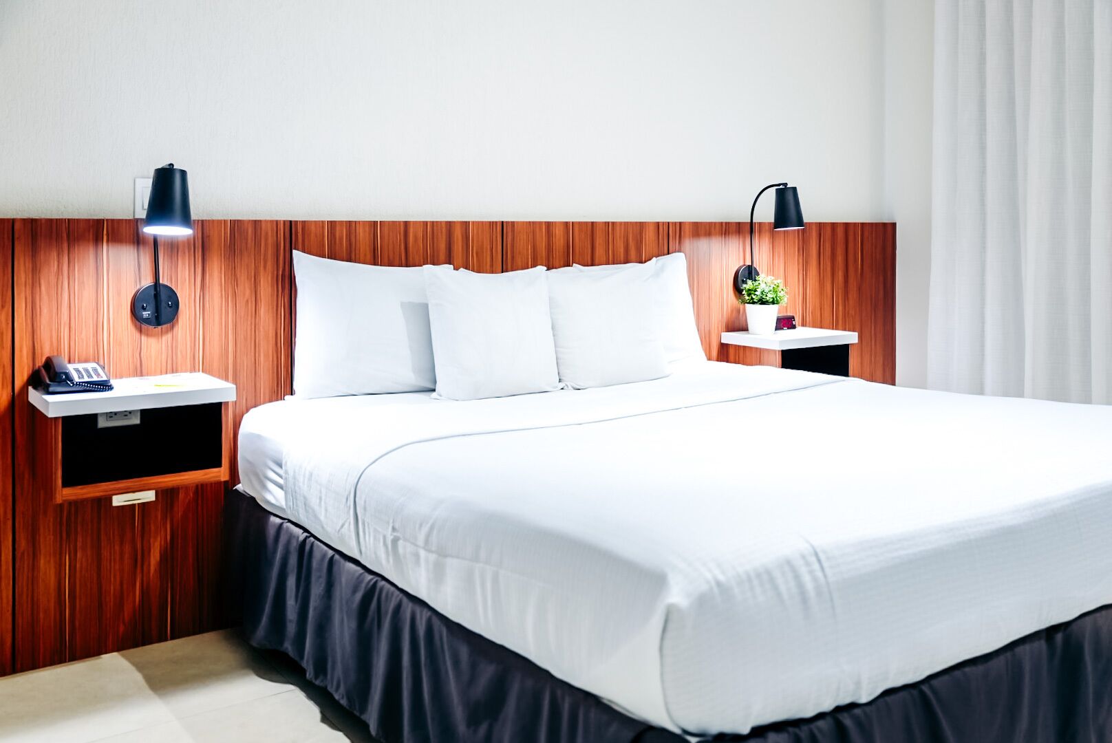 Habitación de hotel con una cama grande, cabecera de madera, mesitas de noche con lámparas, un teléfono y una planta pequeña.