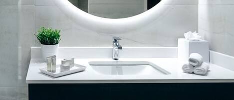 A modern bathroom with a round mirror, white sink, and dark countertop. There is a small plant on the countertop and towels on the shelf below.