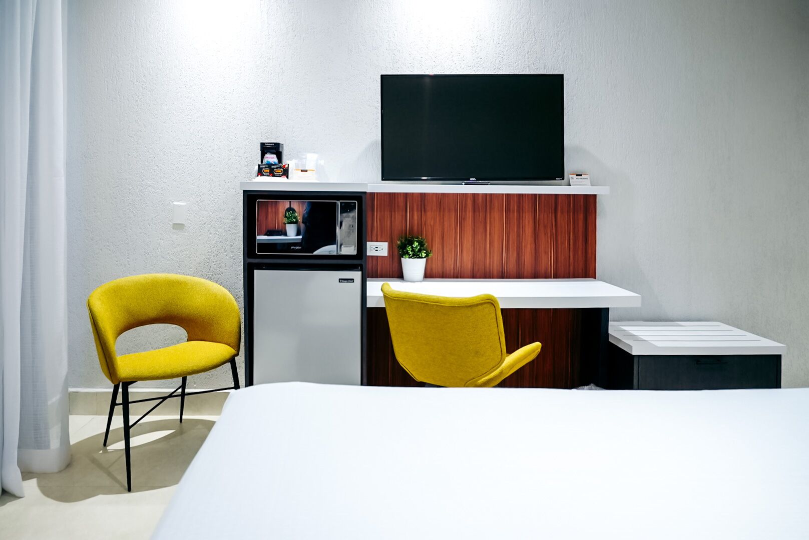 A hotel room with a bed, a desk with a chair, a television, a small refrigerator, and a potted plant.