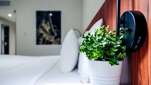 Une chambre d’hôtel avec un lit, du linge de lit blanc, une plante en pot, un appareil fixé au mur et un tableau accroché au mur.