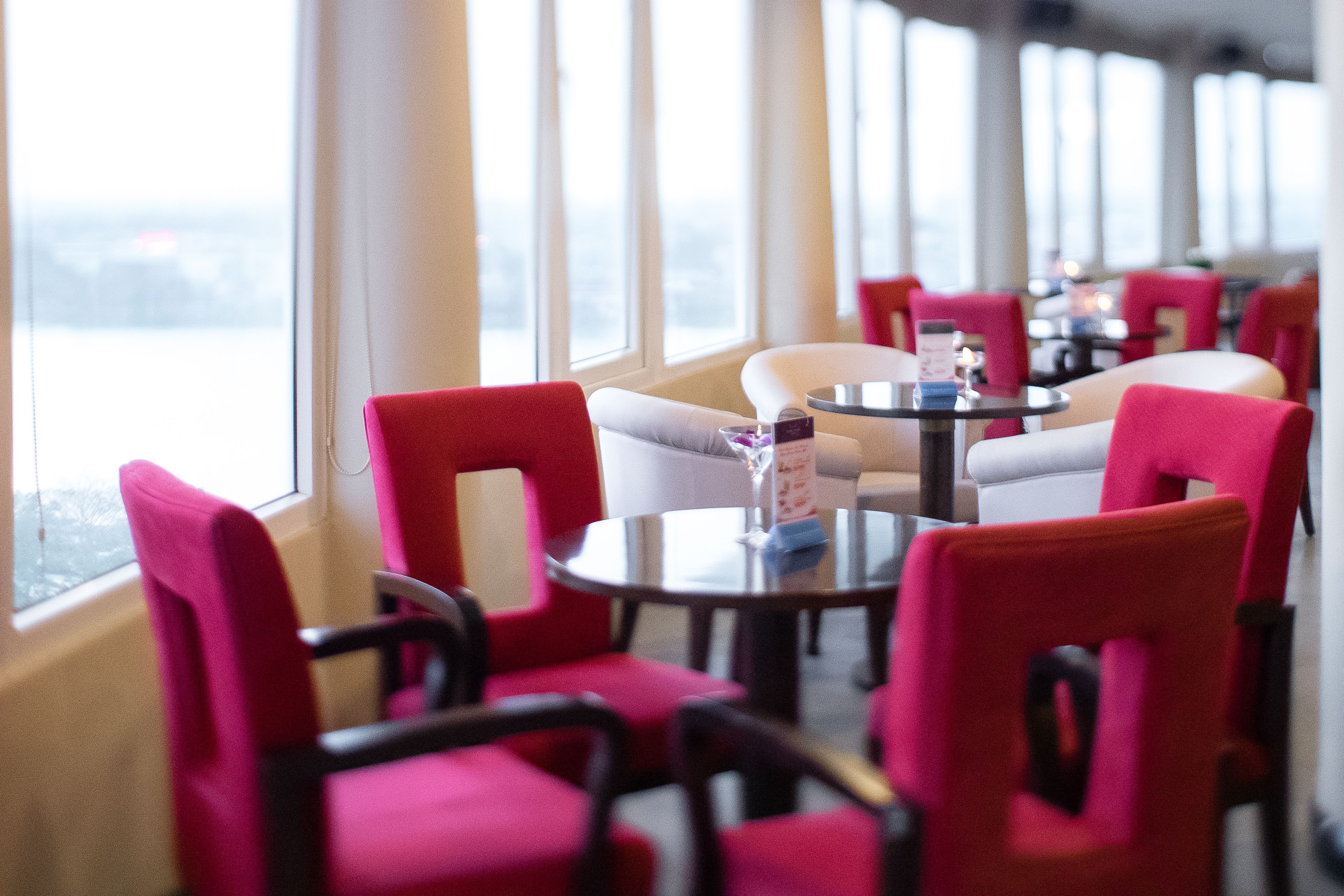 Ein Restaurant mit roten Stühlen und weißen Tischen, großen Fenstern und Blick auf die Stadt.