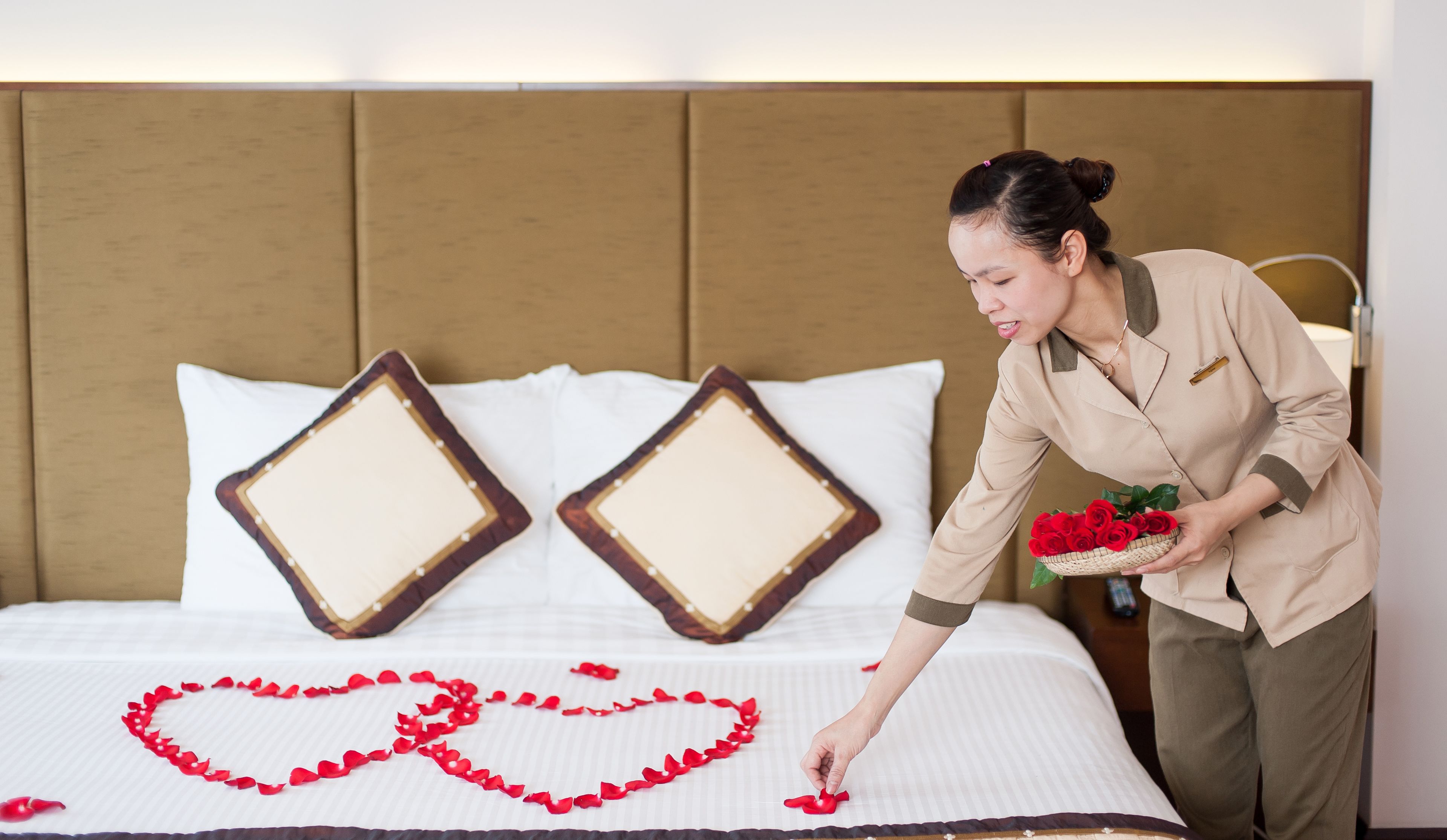 A hotel room with a neatly made bed, two decorative pillows, and a heart-shaped arrangement of rose petals on the bed.