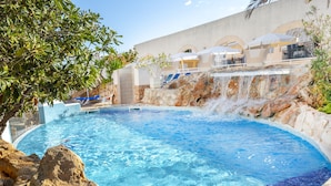 A swimming pool with a waterfall feature, surrounded by a stone wall and a building with outdoor seating. - Hotel Kilimanjaro (Llucmajor)