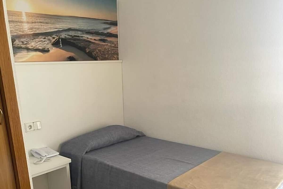 Um quarto pequeno e bem organizado, com uma cama de solteiro, uma mesinha de cabeceira, um telefone fixado na parede e um quadro com uma paisagem de praia.