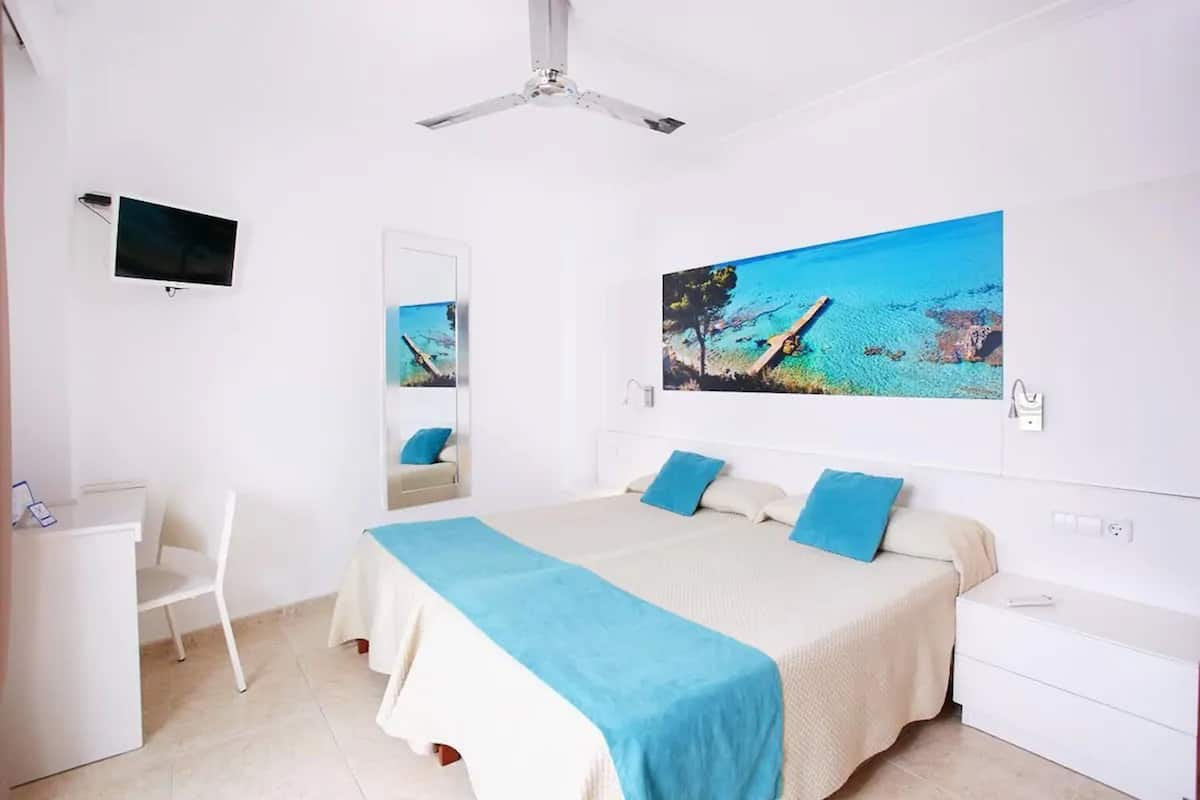 Quarto com cama, escrivaninha, cadeira, ventilador de teto, televisão e uma grande janela com vista para o mar.