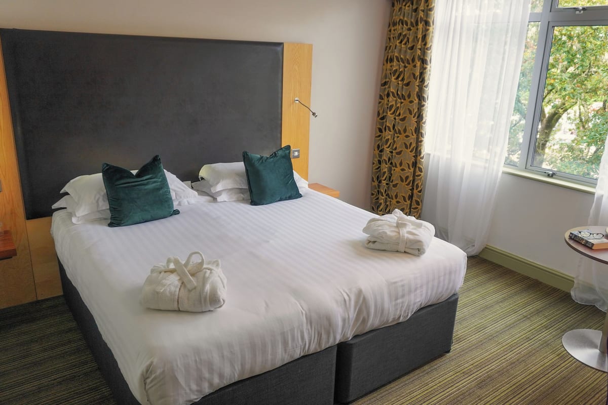 Habitación de hotel con una cama grande, dos almohadas, una mesita de noche con artículos de aseo, una pequeña mesa redonda con fruta y un libro, y una ventana con cortinas.