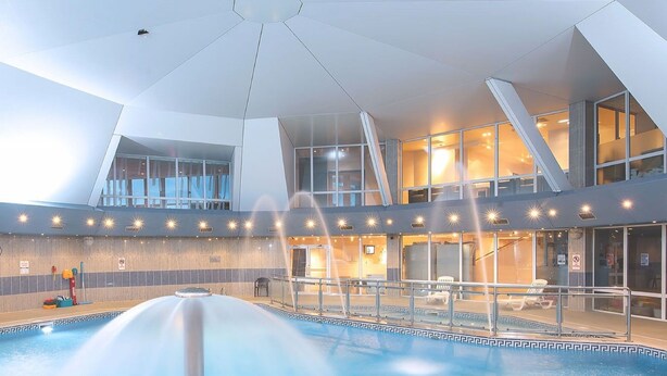 An indoor swimming pool with a central water feature, surrounded by a modern architectural design with large windows and a mezzanine level.