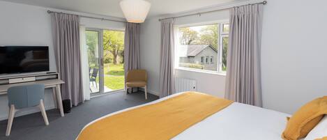 A hotel room with a bed, a desk with a chair, a television, and a view of a house through the window.