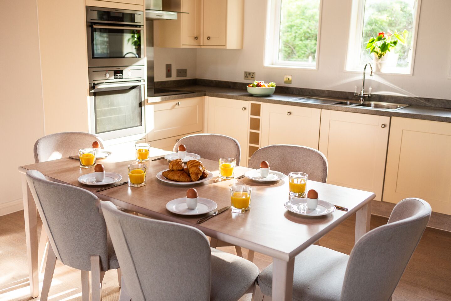 A modern kitchen with a dining table set for breakfast, including croissants, eggs, and orange juice.