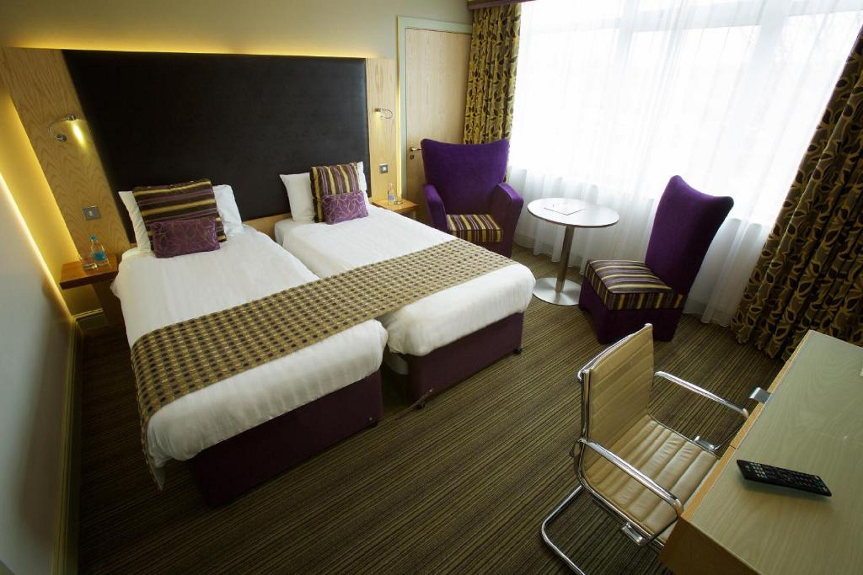 A hotel room with two beds, a small table, a chair, and a desk with a remote control.