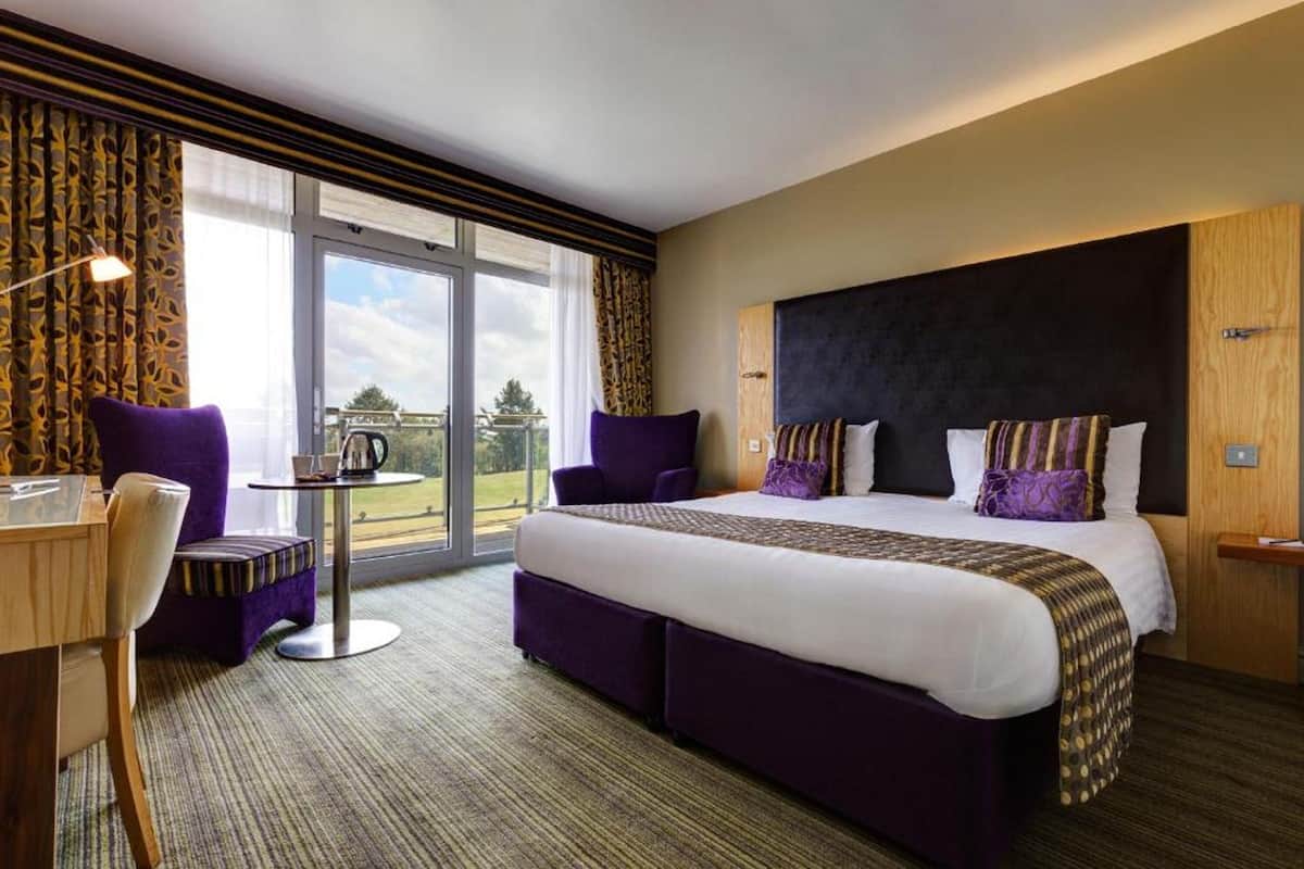 Habitación de hotel con una cama grande, un escritorio y una silla. Hay una ventana con cortinas y vistas al exterior.