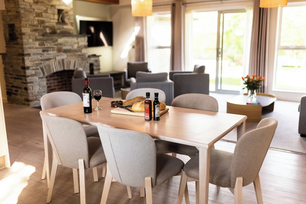 Un comedor con una mesa de madera, sillas tapizadas de gris, una chimenea de piedra y vistas al exterior a través de amplios ventanales.