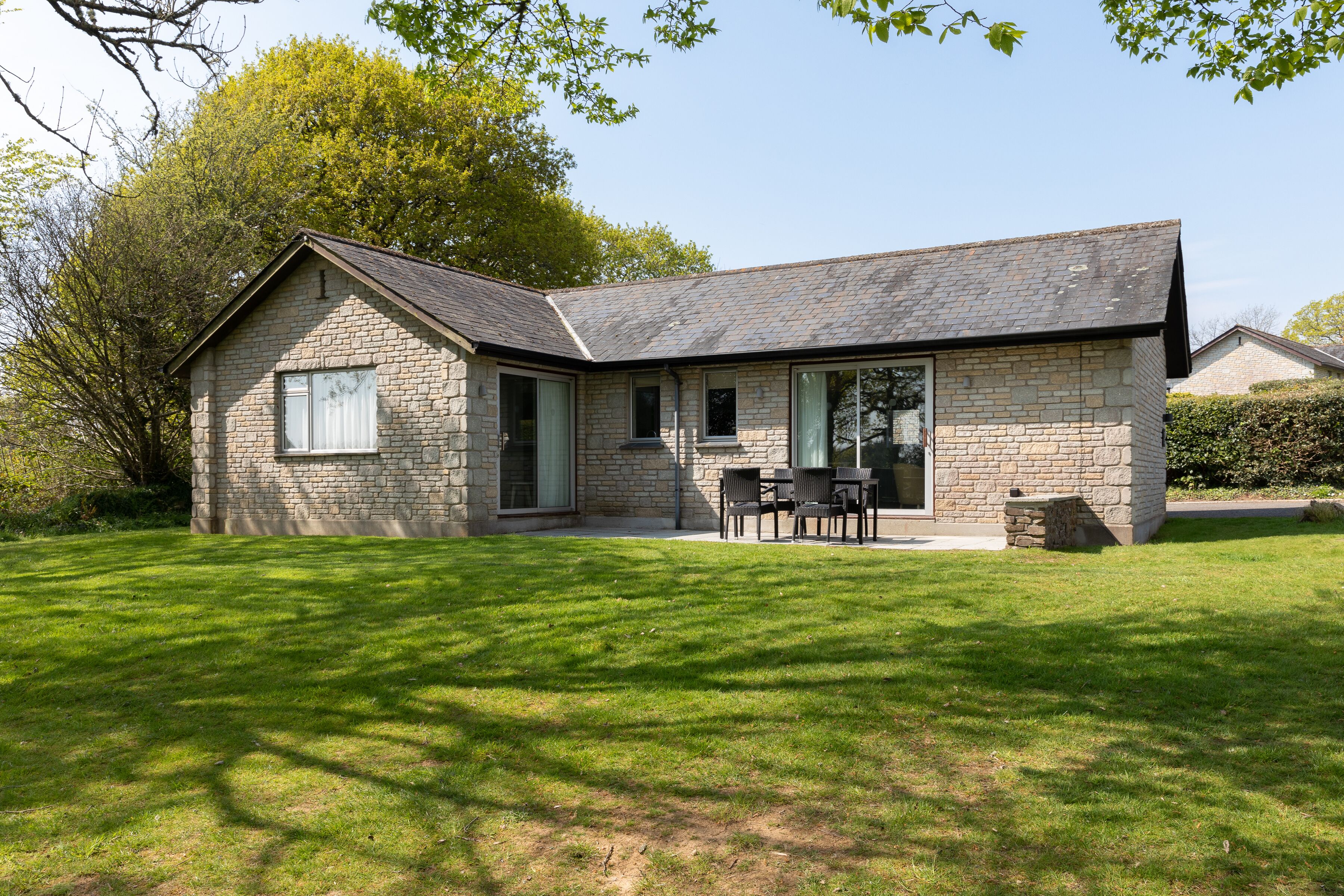 A stone house with a sloped roof, a patio with chairs, and a well-kept lawn.
