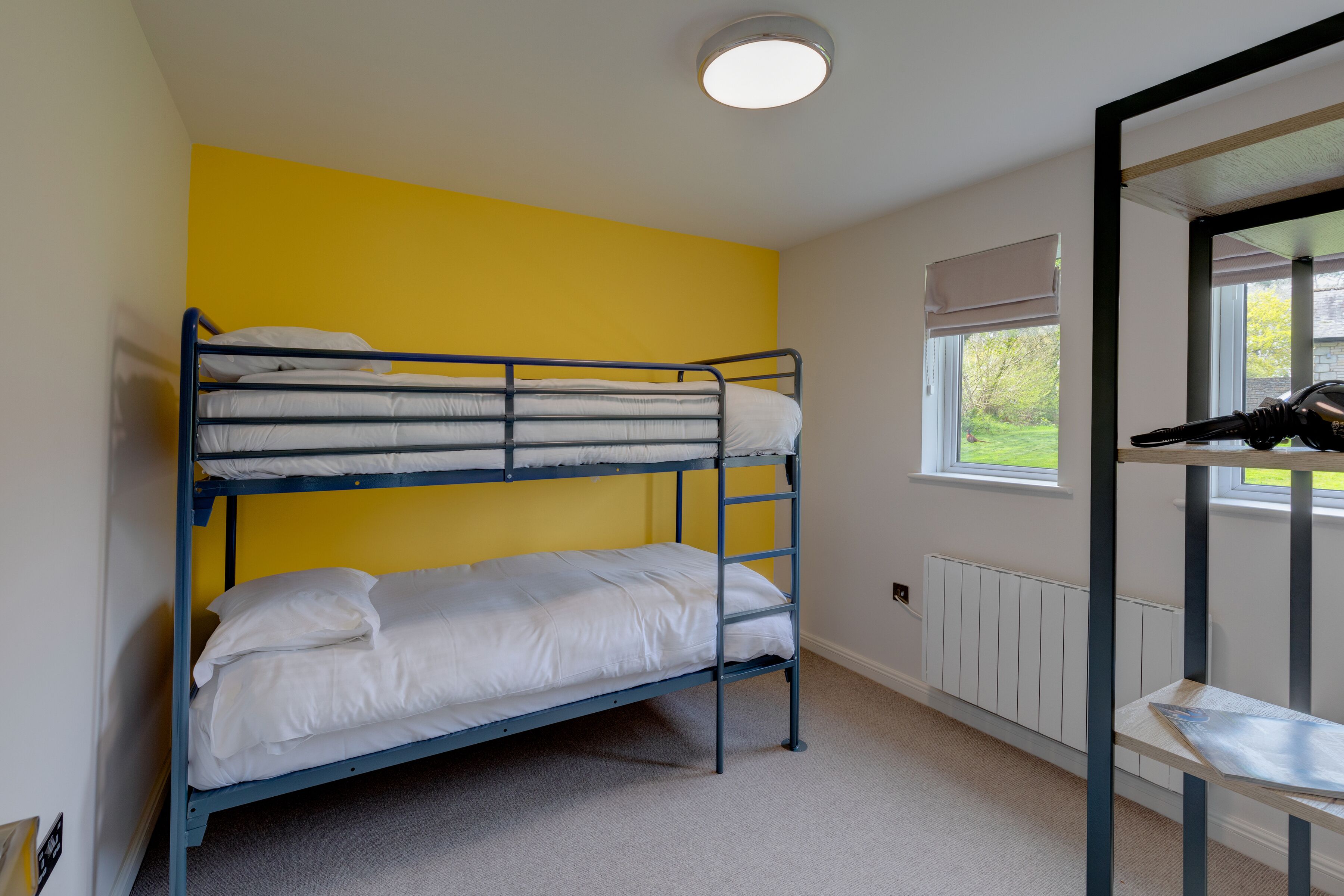 A bunk bed room with a yellow wall, a window with a view of greenery, and a radiator.