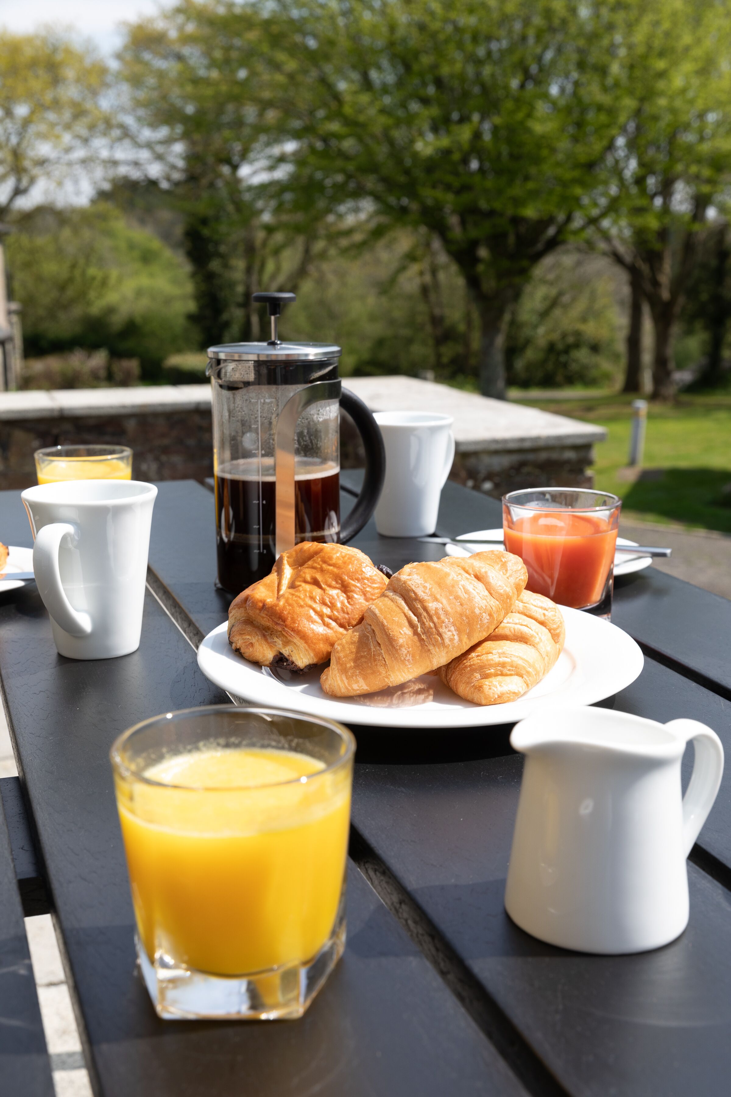 Ein Frühstückstisch im Freien mit Croissants, Saft, Kaffee und einer French Press.
