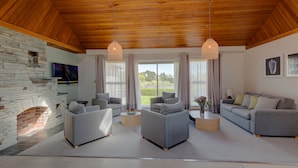 A spacious living room with a stone fireplace, grey sofas, wooden ceiling, and large windows with curtains.
