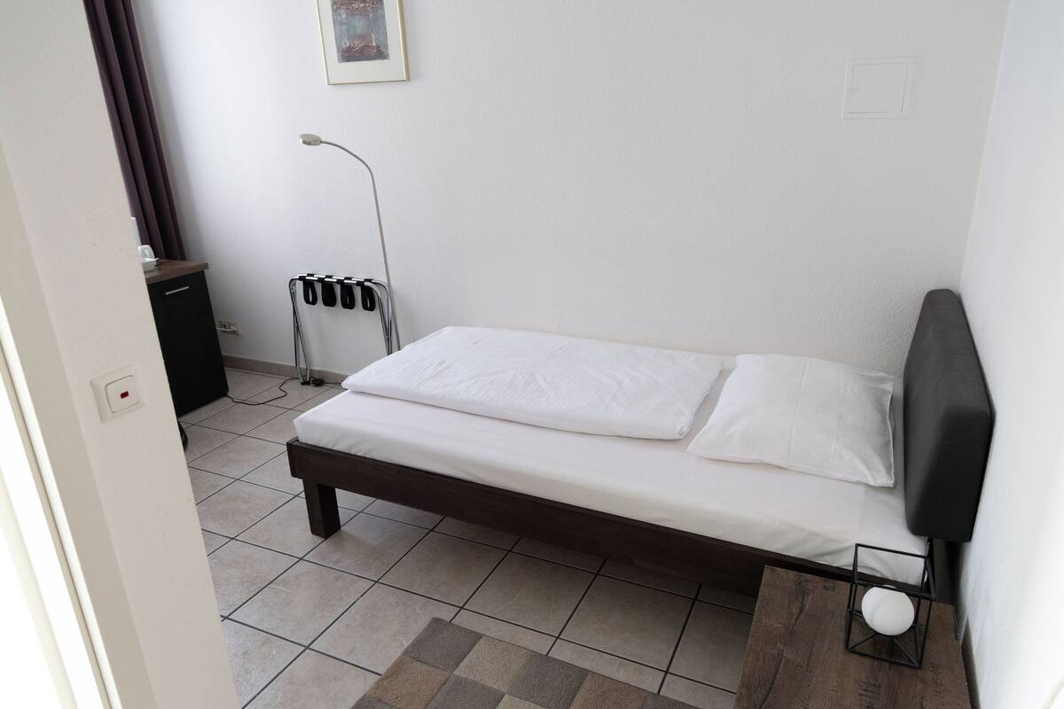A small, neatly arranged bedroom with a single bed, a bedside table, a lamp, and a power outlet.