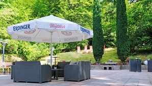 An outdoor seating area with a white umbrella labeled 'ERDINGER Weisbier,' wicker furniture, and a landscaped garden in the background. - Hotel Haus am Park (Bad Homburg v.d. Hoehe)