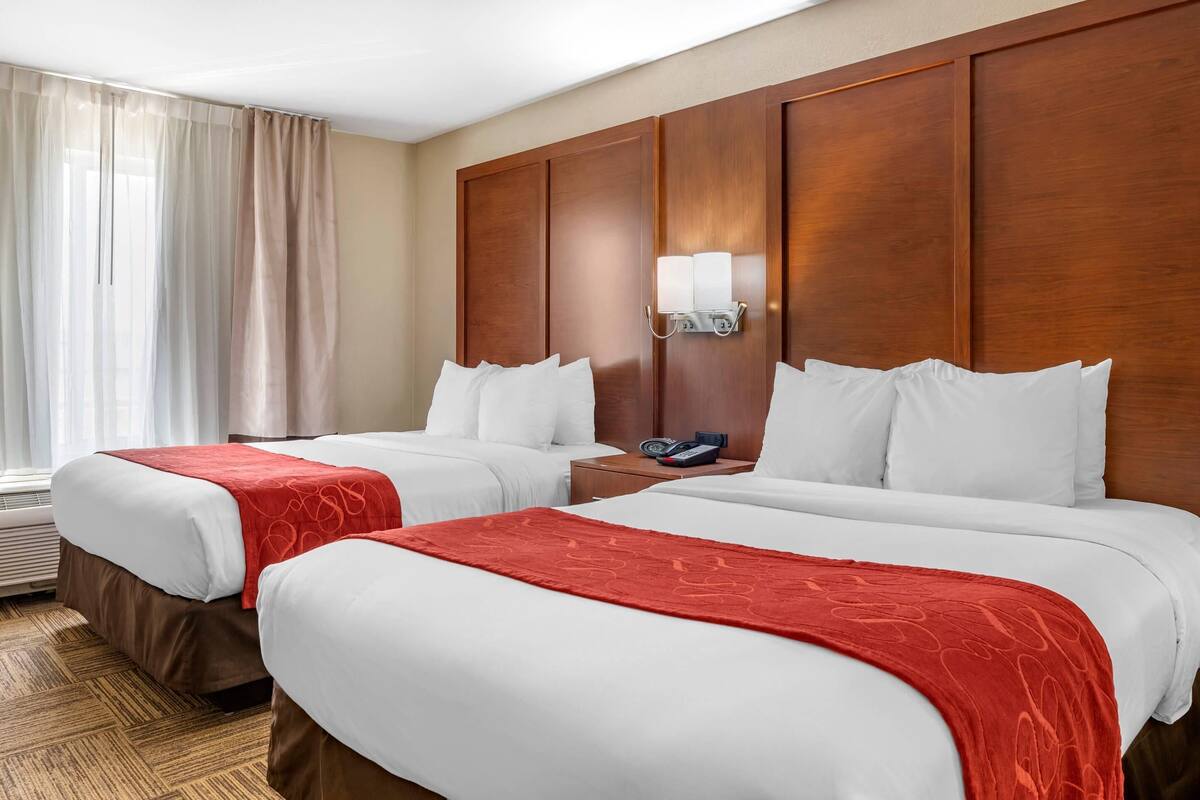Une chambre d’hôtel avec deux lits, une tête de lit en bois, une table de chevet avec un téléphone et une fenêtre avec des rideaux.
