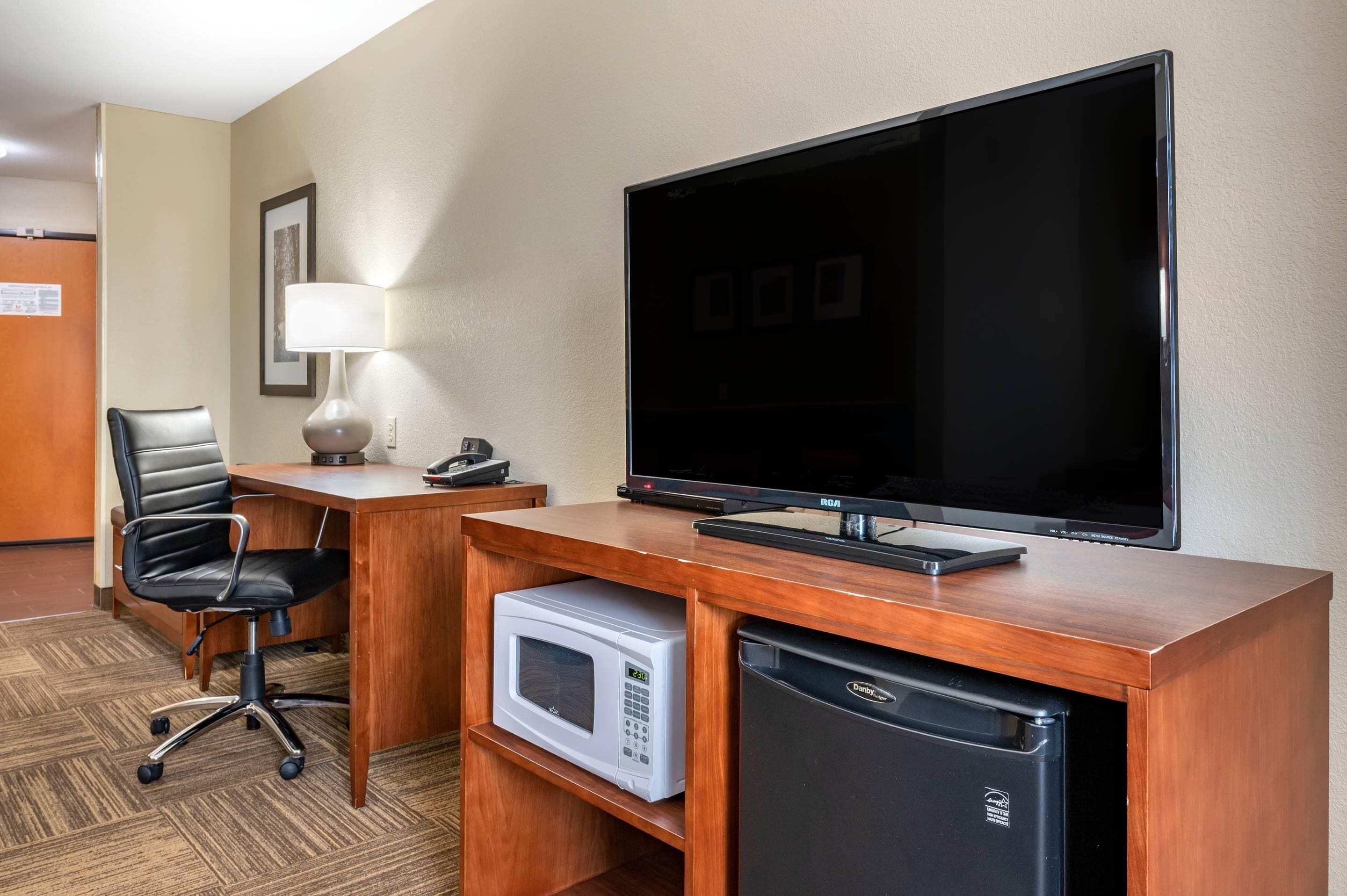 A hotel room with a wooden desk, a black office chair, a flat-screen TV, a microwave, and a mini-fridge.
